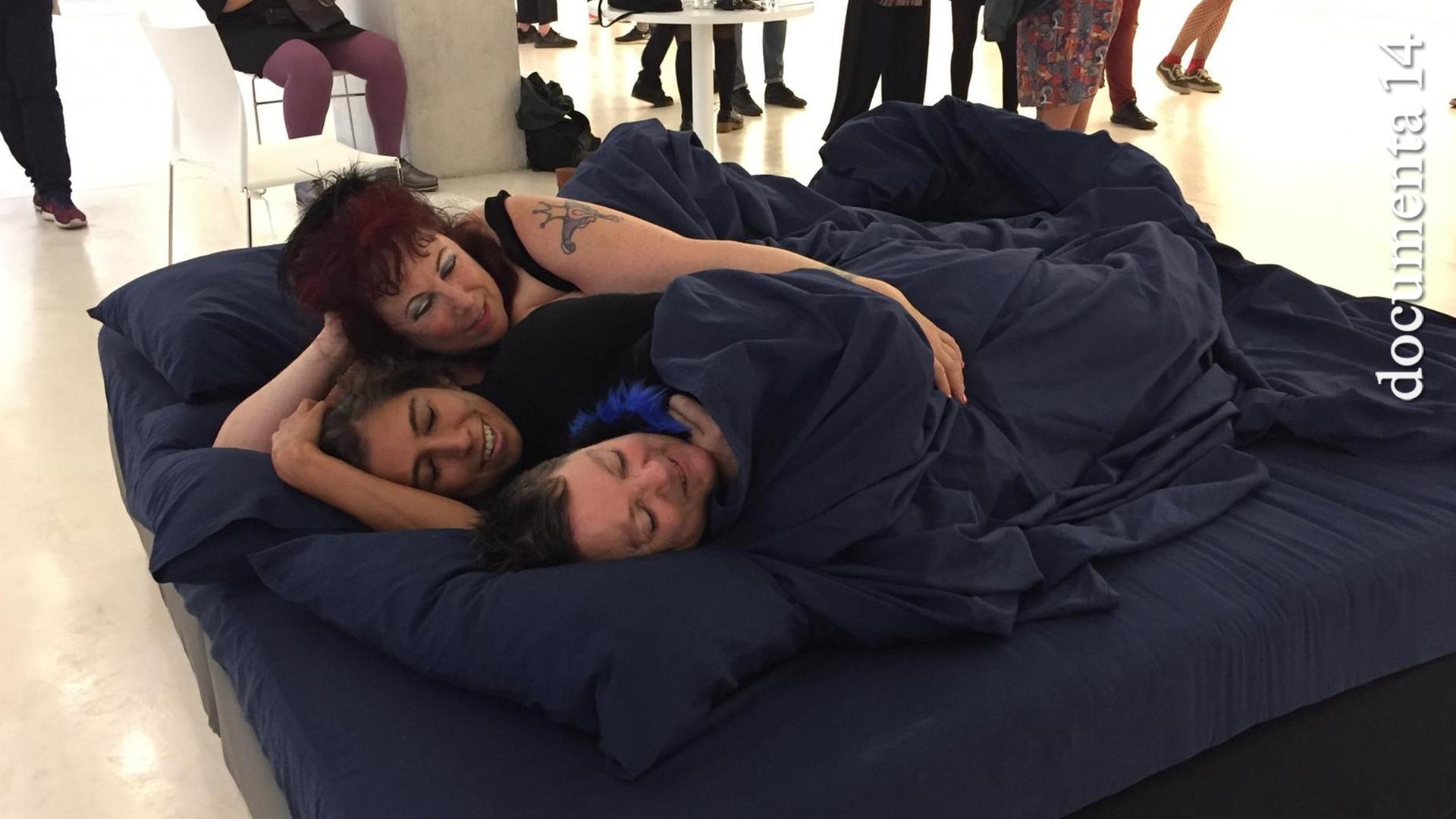 Annie Sprinkle und Beth Stephens haben im Athener Museum für Zeitgenössische Kunst ein Bett aufgebaut und kuscheln darin mit einer Besucherin.
