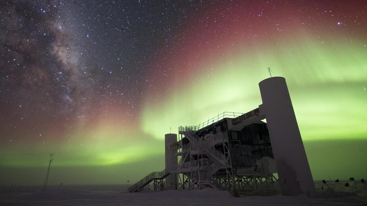 Das IceCube Labor in der Antarktis aus der Froschperspektive. Über der würfelförmigen Station mit zwei Silo-Anbauten leuchtet eine Aurora in Grün und Rot.