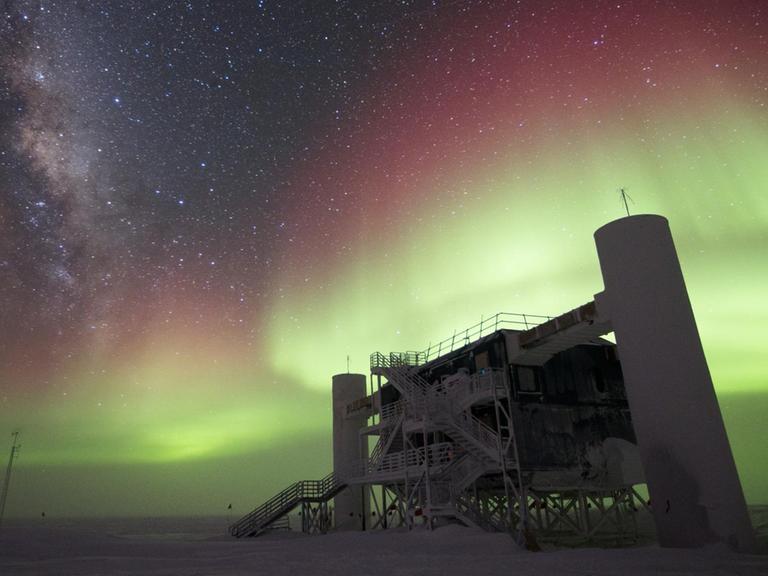 Das IceCube Labor in der Antarktis aus der Froschperspektive. Über der würfelförmigen Station mit zwei Silo-Anbauten leuchtet eine Aurora in Grün und Rot.