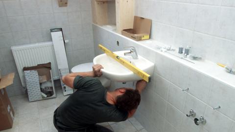 Ein Installateur kniet bei der Montage eines Waschbeckens im Badezimmer auf dem Boden. Mit der Wasserwaage tariert der Sanitärfachmann die korrekte Lage des Beckens aus.