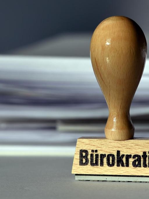 Ein Stempel mit der Aufschrift "Bürokratie" steht vor einem Stapel Papier.