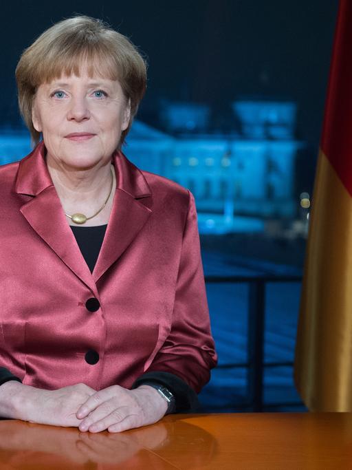 Angela Merkel an einem Schreibtisch vor Deutschland- und EU-Flagge, im Hintergrund der Blick durchs Fenster auf den Reichstag.