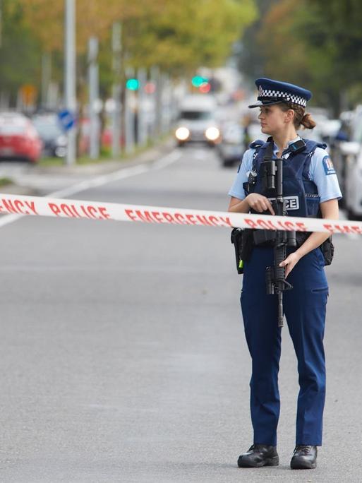 Eine Polizistin trägt ein Gewehr und steht hinter einer Polizeiabsperrung auf einer Straße.