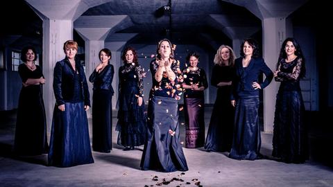 Das Ensemble Tiburtina steht in einem Halbkreis in einem Keller-Gewölbe, die 9 Sängerinnen tragen schwarze Konzertkleidung. Die Leiterin vorne in der Mitte, Barbora Kabátková, pustet bunte Blätter in die Luft