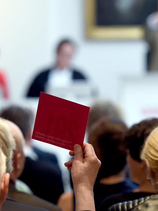 Köpfe von Auktionsteilnehmer von hinten fotografiert, ein Mann hält eine rote Karte hoch, die Auktionatoren im Hintergrund sind verschwommen.