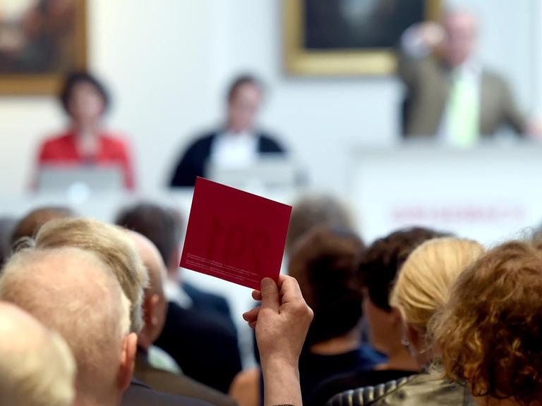 Köpfe von Auktionsteilnehmer von hinten fotografiert, ein Mann hält eine rote Karte hoch, die Auktionatoren im Hintergrund sind verschwommen.