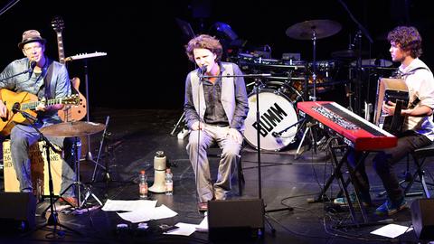 Felix Meyer sitzt singend auf der Bühne des Theaterkahns in Dresden zwischen dem Gitarristen und dem Keyboarder, der gerade auf einem Akkordeon spielt