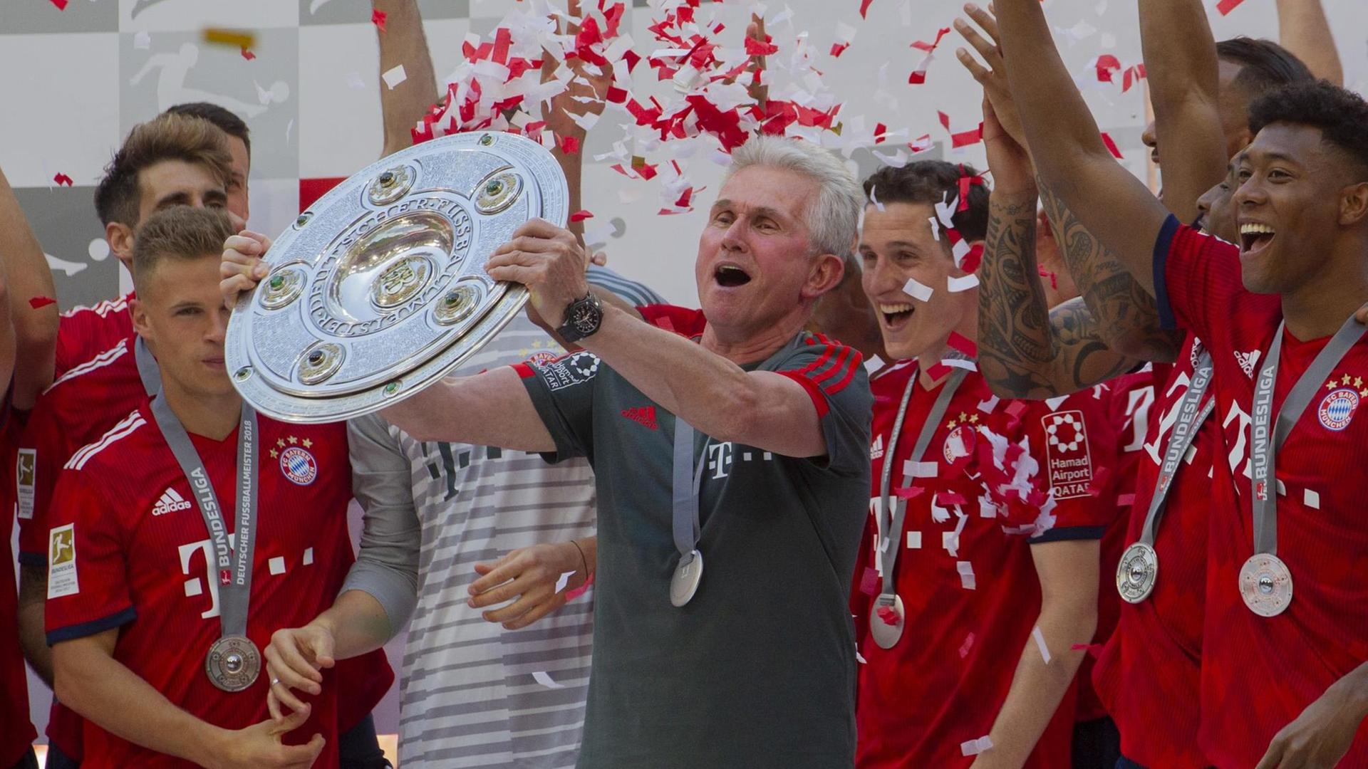 Jupp Heynckes steht zwischen den Spielern Joshua Kimmich, Sebastian Rudy und David Alaba und hält die Meisterschale hoch, die der Verein in der Saison 2017/18 gewonnen hat. Im Hintergrund rieseln rote und weiße Papierfetzen herab, die Farben des Klubs.