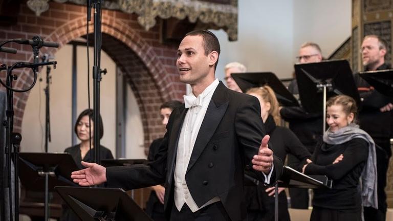 Der Kantor singt in einer Kirche gemeinsam mit einem Chor, der hinter ihm steht.