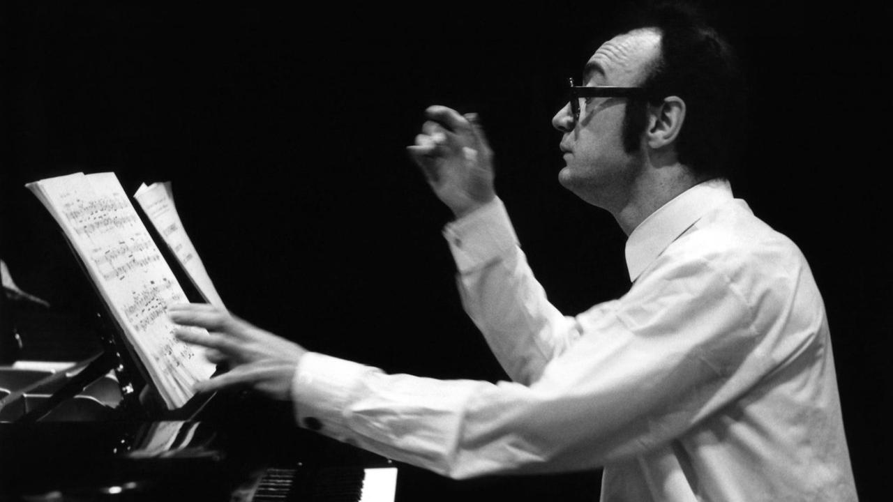 Schwarz-Weiß Fotografie von 1971, der Pianist Alfred Brendel sitzt am Flügel (links) im Herkulessaal in München, die linke Hand liegt auf den Noten vor ihm auf dem Pult, die Rechte scheint zu dirigieren. Er trägt ein weißen Hemd und eine dunkle Hornbrillle.