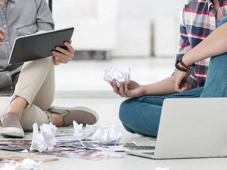Zwei junge Menschen sitzen mit Laptops auf dem Boden - vor ihnen zerknülltes Papier.