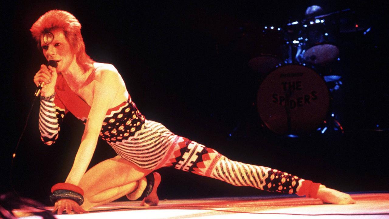 David Bowie kniet in einem auffälligen Kostüm auf einer Bühne.