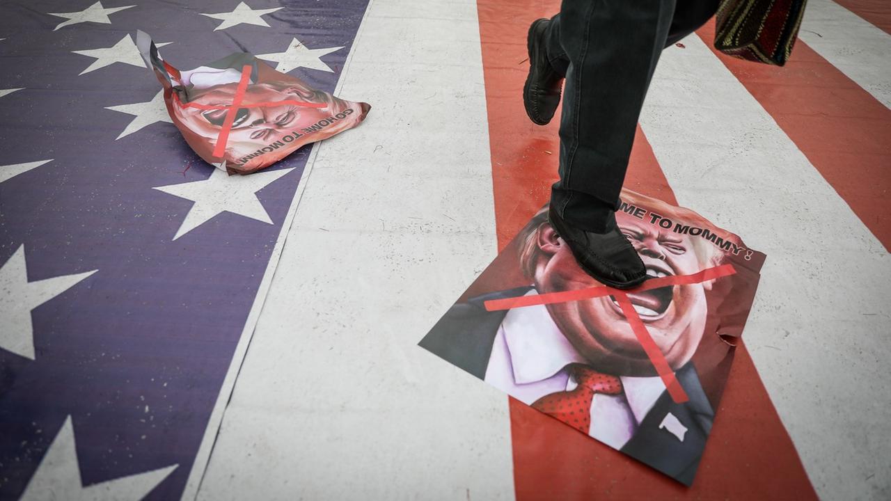 Plakate mit einer Donald Trump Karikatur liegen auf dem Boden eine Bürgersteigs in Teheran, der mit der amerikanischen Flagge bemalt ist. Darauf läuft eine Person, wobei das Bild am Rumpf endet.