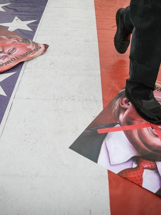 Plakate mit einer Donald Trump Karikatur liegen auf dem Boden eine Bürgersteigs in Teheran, der mit der amerikanischen Flagge bemalt ist. Darauf läuft eine Person, wobei das Bild am Rumpf endet.