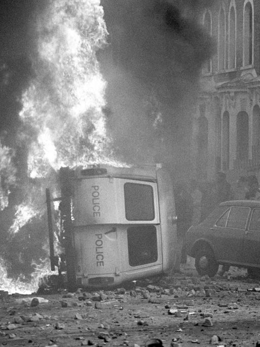 Ein Kleinbus der Polizei liegt auf der Seite und brennt. Die Straße ist übersäht mit geworfenen Steinen.