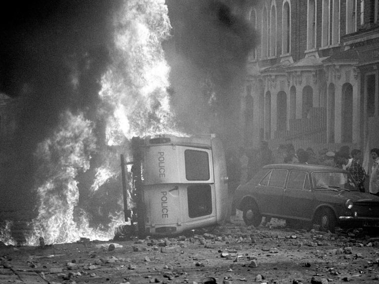 Ein Kleinbus der Polizei liegt auf der Seite und brennt. Die Straße ist übersäht mit geworfenen Steinen.