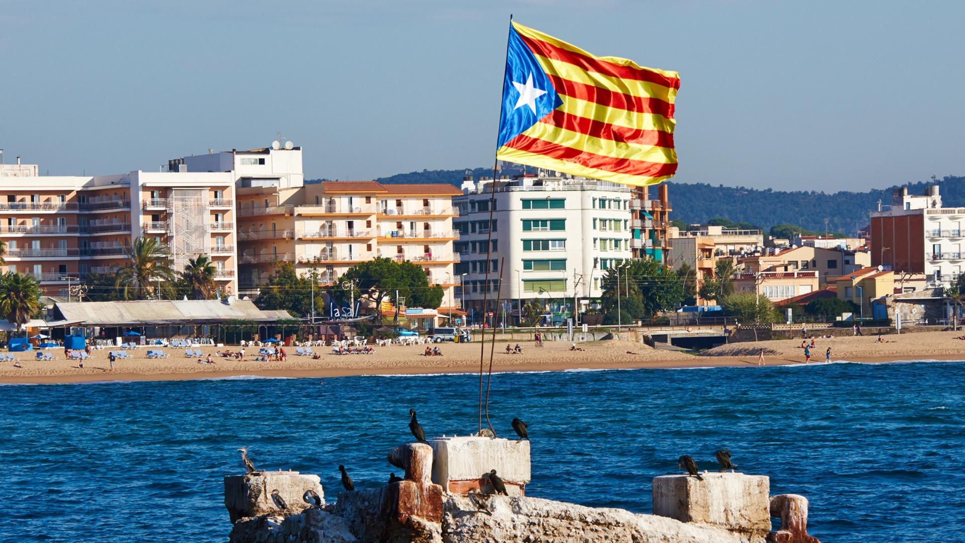 Ein Felsen im Meer vor der Küste Kataloniens, auf der eine katalanische Flagge im Wind weht.