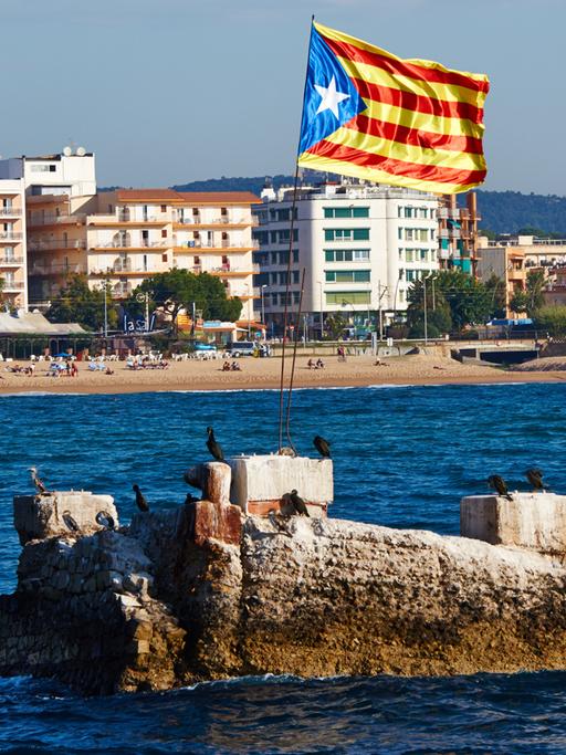 Ein Felsen im Meer vor der Küste Kataloniens, auf der eine katalanische Flagge im Wind weht.