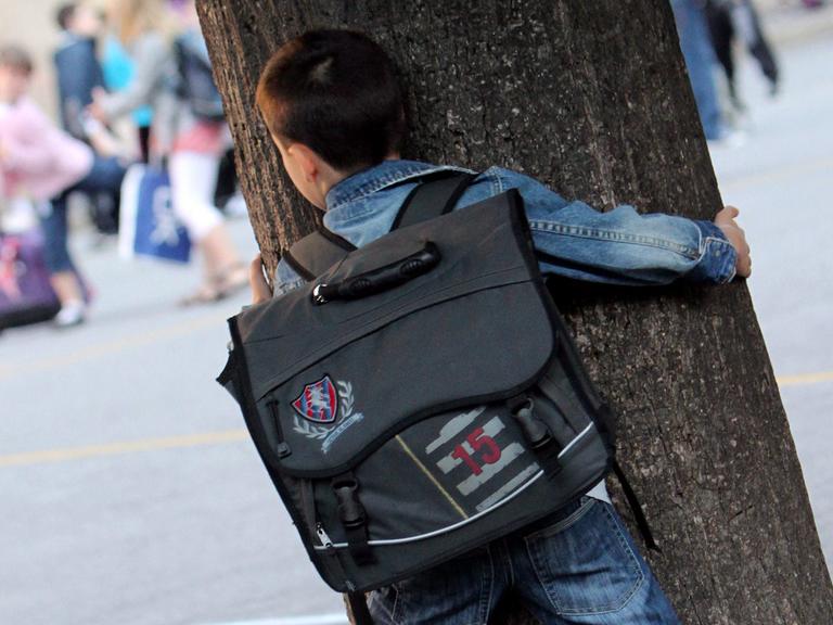 Junge mit Tornister auf dem Rücken, der sich auf dem Schulhof hinter einem Baum versteckt.