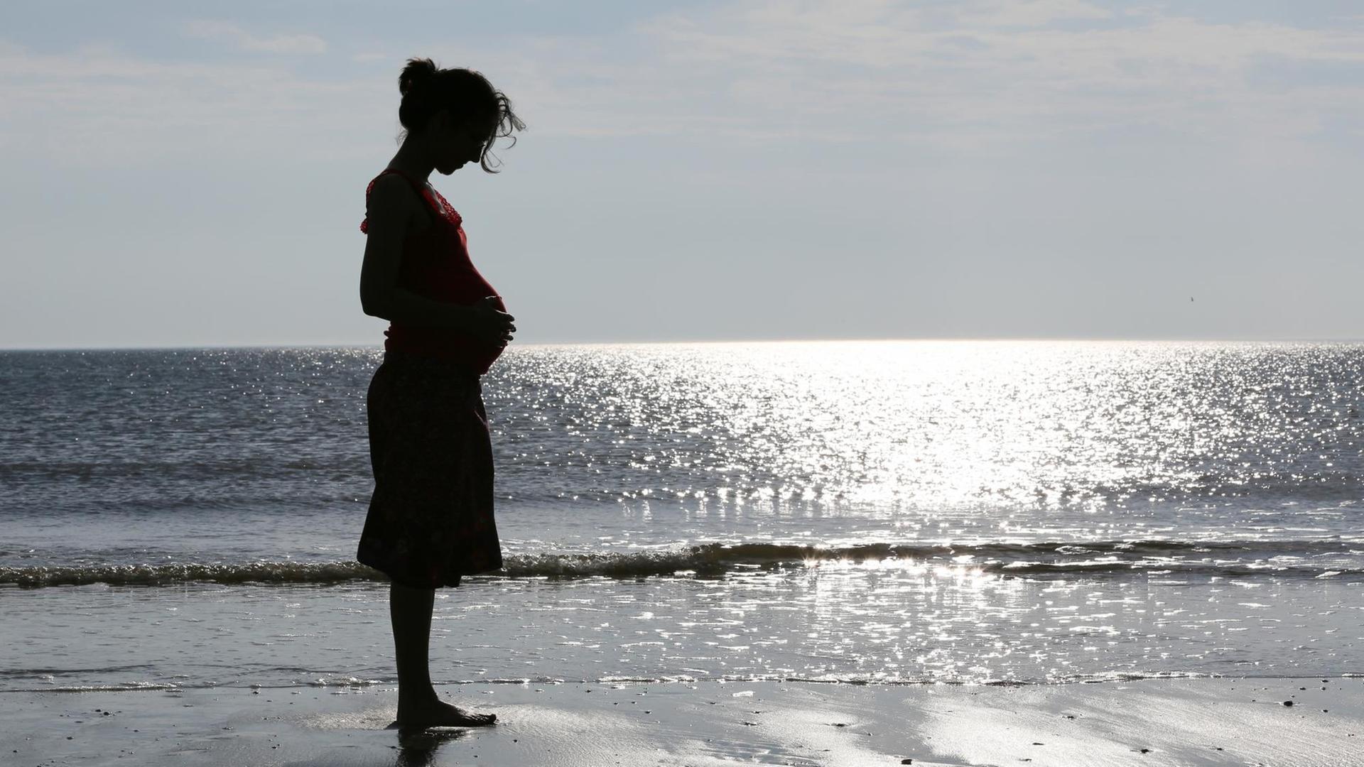 Eine schwangere Frau steht am Meer und hält sich den Bauch 