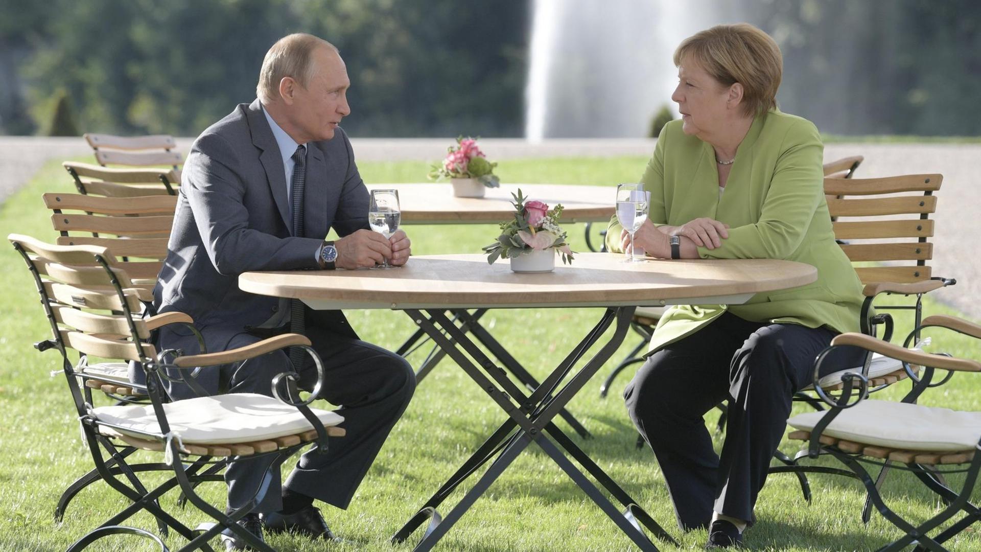 Putin und Merkel sitzen auf Garten-Stühlen in dem Park von Schloss Meseberg.