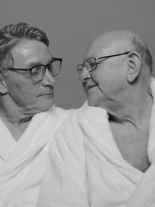 Zwei ältere Männer in weißen Bademänteln sitzen zusammen auf einer Couch und schauen sich gegenseitig an.