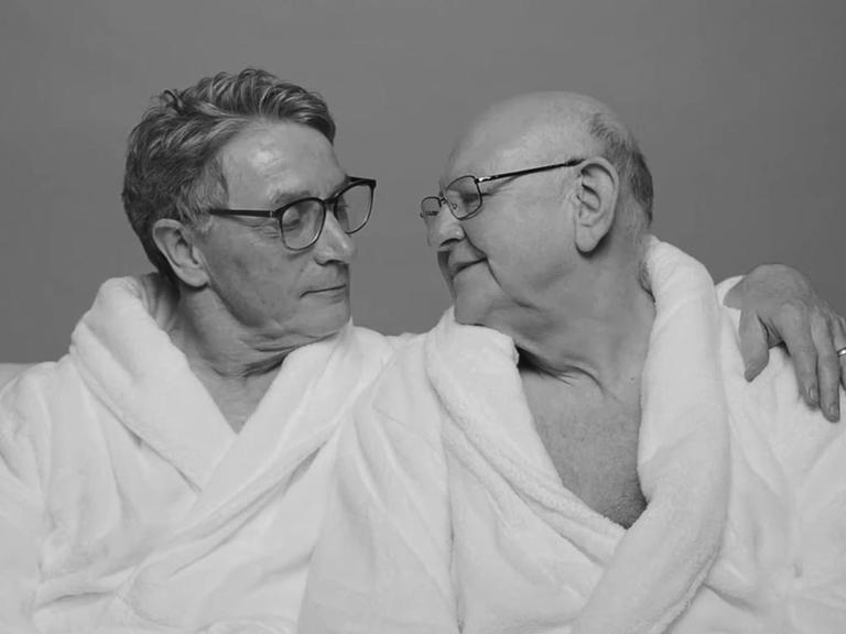 Zwei ältere Männer in weißen Bademänteln sitzen zusammen auf einer Couch und schauen sich gegenseitig an.