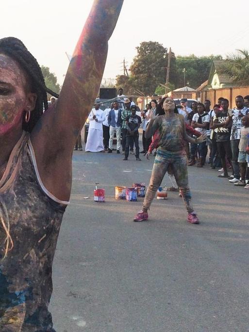 Straßenperformance der kongolesischen Künstlerin Sarah Mukadi. Sie ist am ganzen Körper bunt bemalt und hebt die Arme.