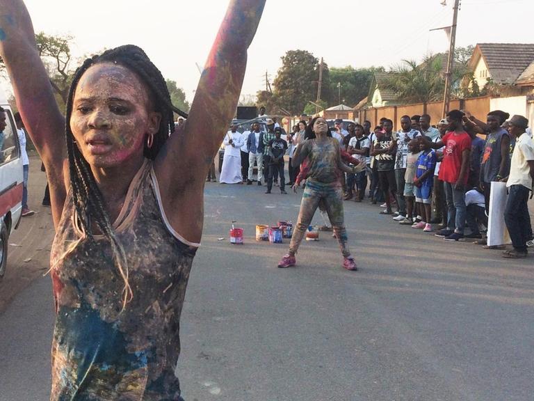 Straßenperformance der kongolesischen Künstlerin Sarah Mukadi. Sie ist am ganzen Körper bunt bemalt und hebt die Arme.