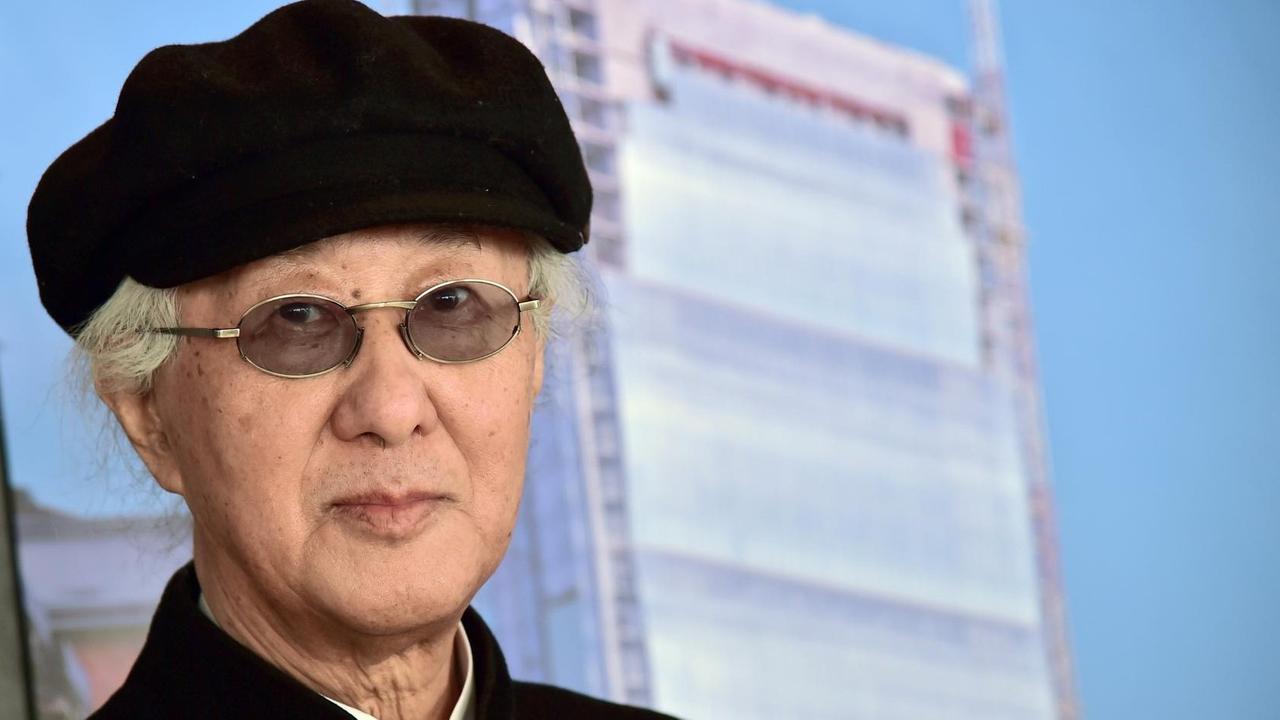 Das Bild zeigt den japanischen Architekten Arata Isozaki. Er trägt eine schwarze Stoffmütze. Im Hintergrund ist ein gläsernes Hochhaus zu sehen.