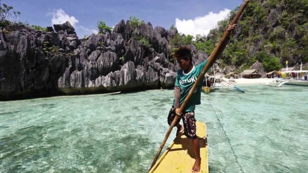Ein Mann steht auf einem Surfbrett im Meer. Im Hintergrund ist Coron, eine Stadtgemeinde die zur Provinz Palawan gehört, zu sehen.
