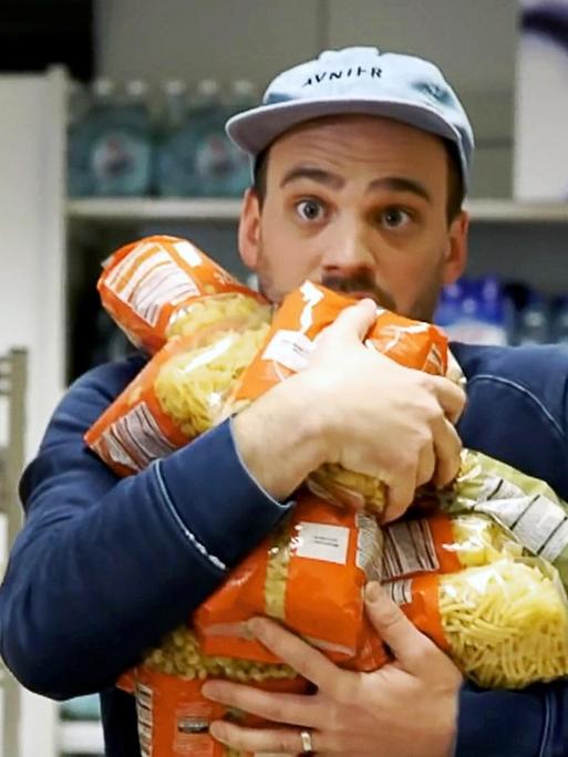 Ein junger Mann im Supermarkt versucht so viele Pasta Packungen wie möglich in seinen Armen zu trasportieren.