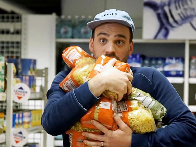 Ein junger Mann im Supermarkt versucht so viele Pasta Packungen wie möglich in seinen Armen zu trasportieren.