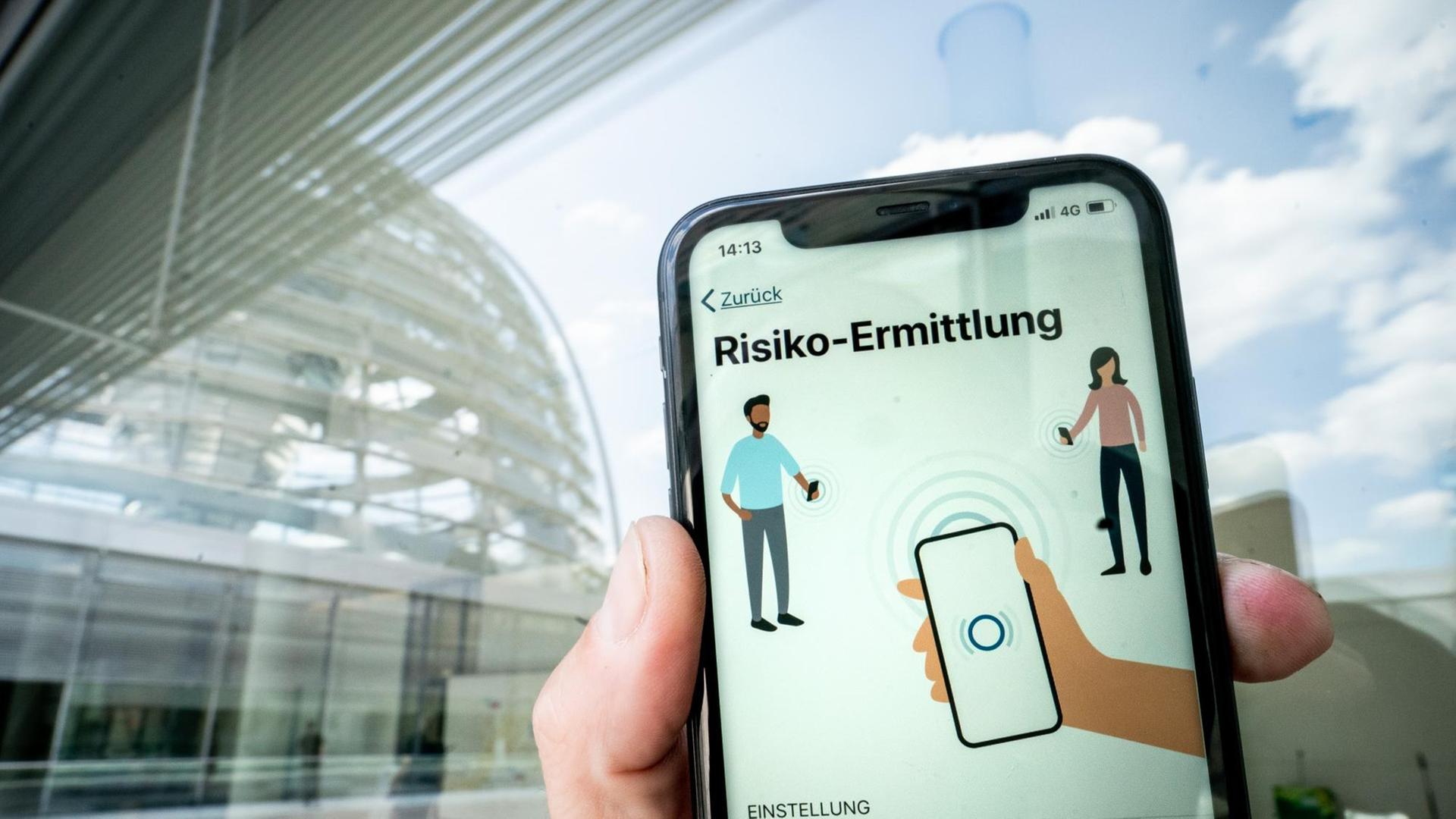 Die Corona-Warn-App mit der Seite zur Risiko-Ermittlung ist im Display eines Smartphone vor der Kuppel des Reichstags zu sehen.