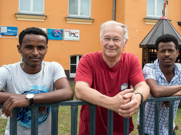 Der Bundestagsabgeordnete Martin Patzelt (CDU) und die beiden Flüchtlinge aus Eritrea Haben und Awet am Zaun des Gemeinde- und Vereinshauses in Briesen im Landkreis Oder-Spree (Brandenburg). Sie lehnen sich über ein Geländer und schauen in die Kamera.