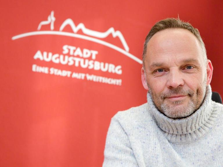 Dirk Neubauer vor einem roten Hintergrund, auf dem Stadt Augustusburg steht. Er trägt einen hellen Rollkragenpullover und hat einen Vollbart.