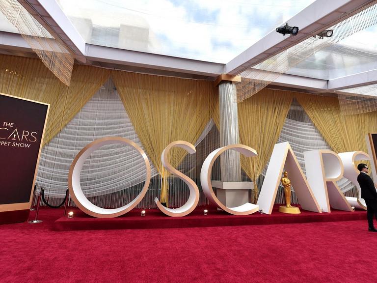 Blick auf den "Oscars"-Schriftzug, der 2020 in einem Raum auf einem roten Teppich stand. Daneben Schilder mit der Aufschrift "The Oscars Red Carpet Show", eine Vorabshow vor der Verleihung der Oscars. Am Rand stehen in Abendrobe gekleidete Personen.