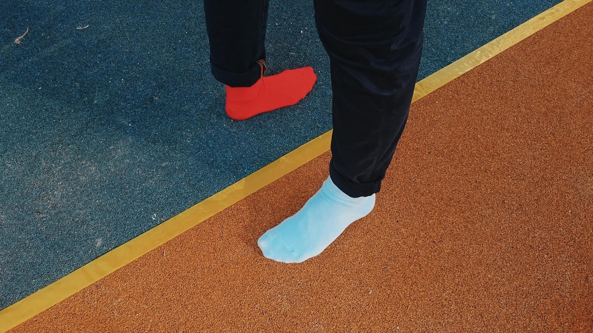 Auf einer Sportlaufbahn steh ein Fuß in einem blauen Socken und ein Fuß in einem orangenem Socken in unterschiedlichen Richtungen.