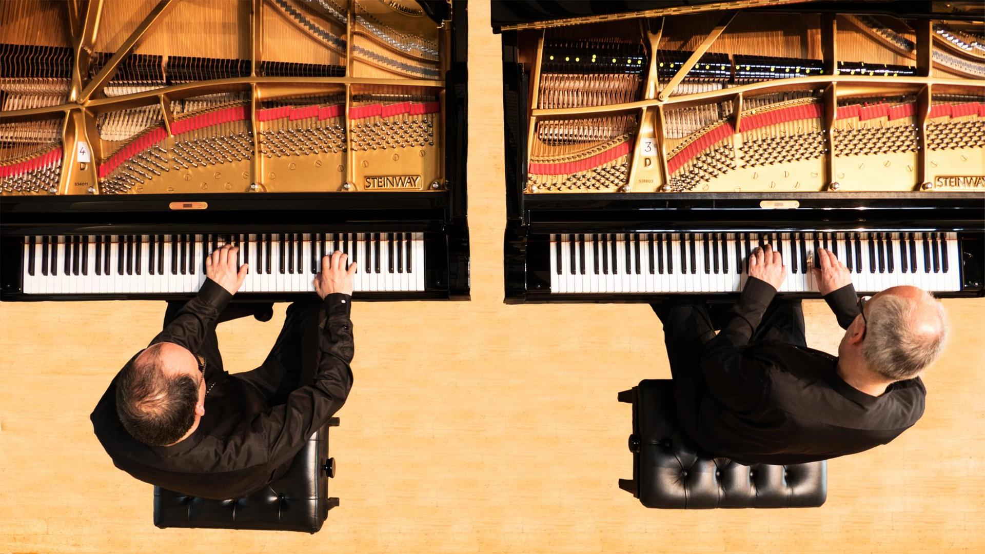 Aufsicht auf zwei Herren die an jeweils einem Konzertflügel sitzen, die Hände auf der Tastaur. Sie tragen schwarze Konzertkleidung, sitzen auf schwarzen Klavierhockern, unter ihnen sieht man hellen Holzboden.