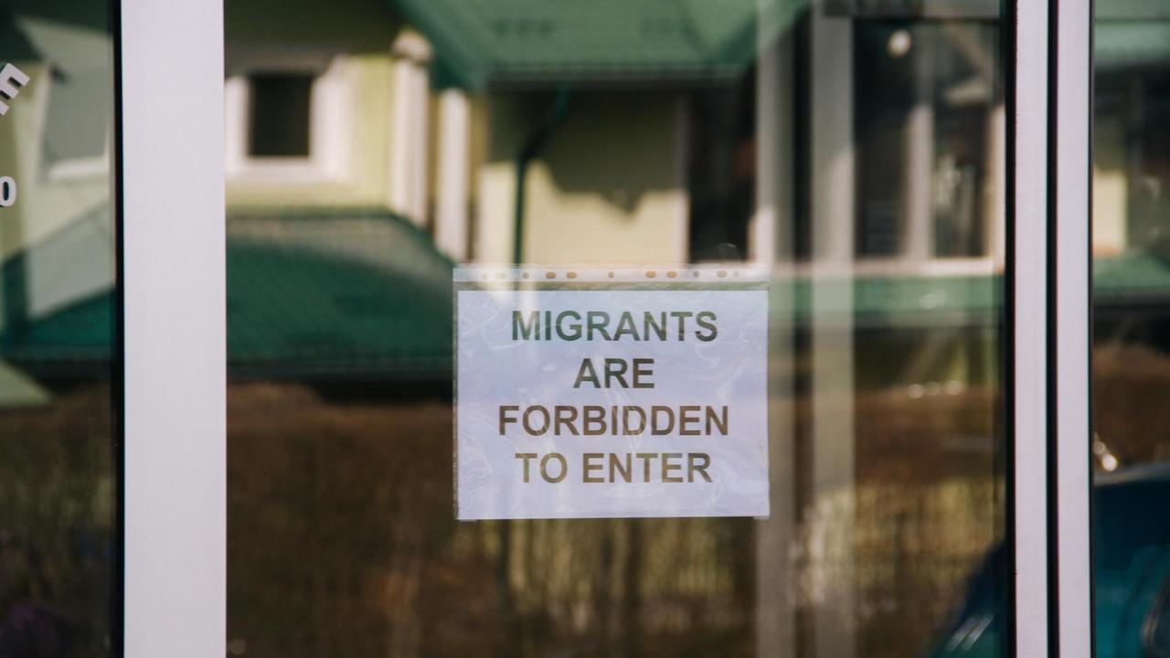 An einer Glastür zu einem Supermarkt ist ein Schild angebracht, auf dem steht "Migrants are forbidden to enter" - Migranten dürfen hier nicht hinein.