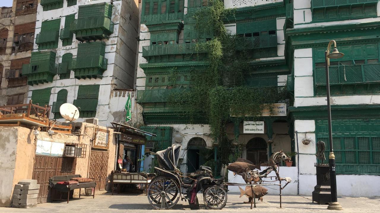 In der Altstadt von Jeddah, vor Häusern mit den typischen grünen Holzvorbauten vor den Fenstern, steht eine Skulptur aus rostigem Metall in Gestalt einer Kutsche mit Pferd, flankiert von zwei großen Fischen.