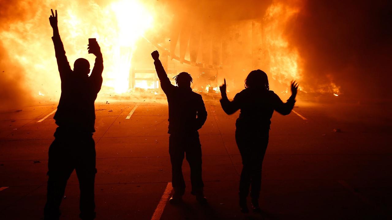 Drei Protestierende auf der Straße, im Hintergrund brennt es. 