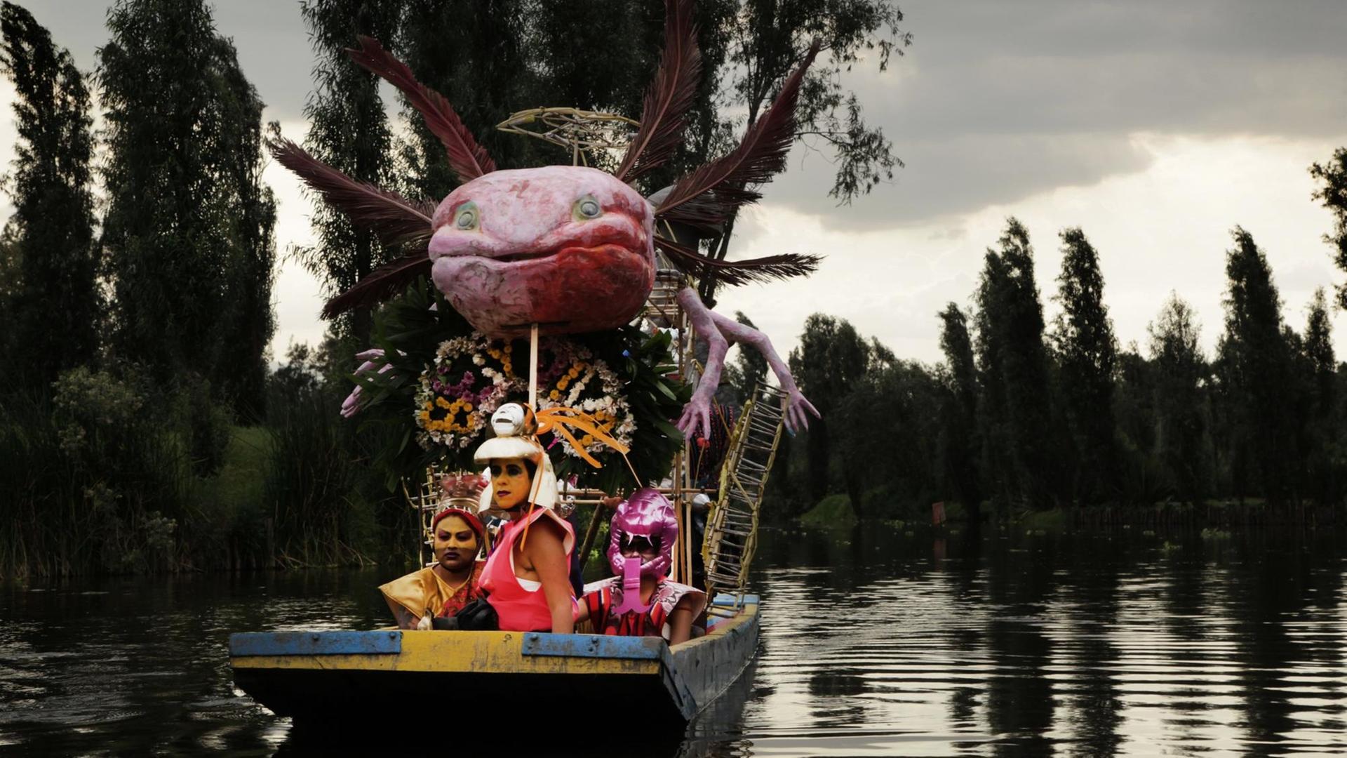 Drei fantasievoll verkleidete Menschen sitzen in einem Boot, auch dieses ist reichlich geschmückt. Darin thront auf einem Gerüst eine überdimensionierte Nachbildung des mexikanischen Schwanzlurchs Axolotl mit Armen und Federn.