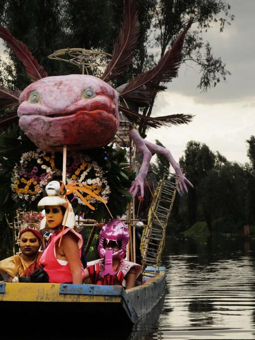 Drei fantasievoll verkleidete Menschen sitzen in einem Boot, auch dieses ist reichlich geschmückt. Darin thront auf einem Gerüst eine überdimensionierte Nachbildung des mexikanischen Schwanzlurchs Axolotl mit Armen und Federn.