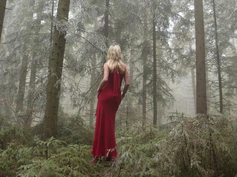 Eine Frau in rotem Abendkleid steht in einem undurchdringlichen Wald und wendet uns den Rücken zu.