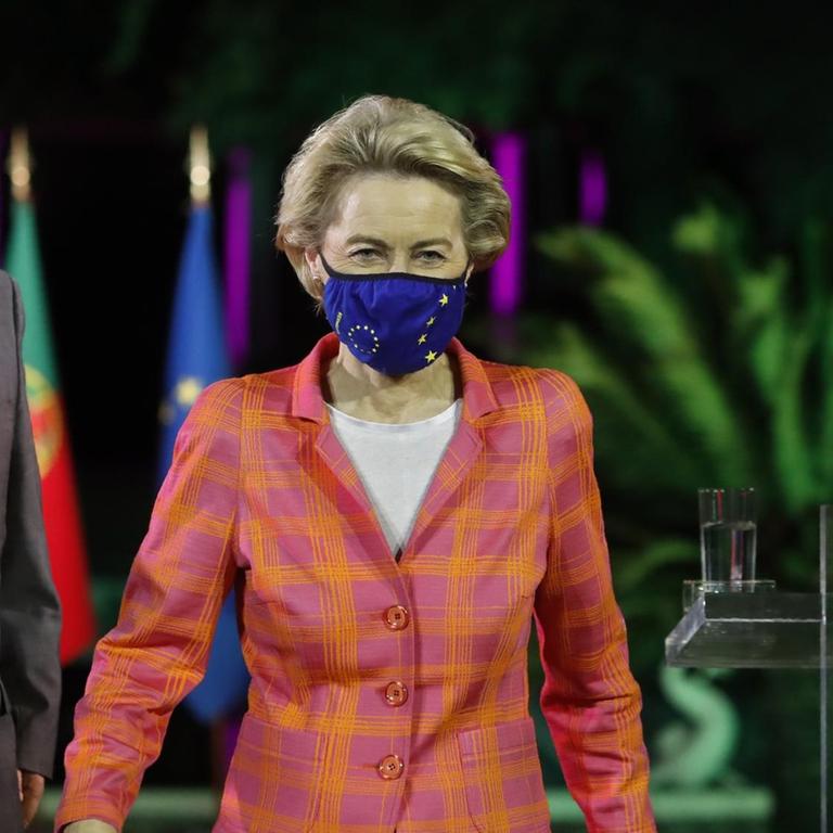 Die beiden gehen hintereinander zu einem Rednerpult aus Plexiglas, beide tragen einen Mund-Nasen-Schutz. Von der Leyen im orangefarbenen Jackett geht voraus. 