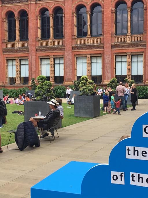 Alteingesessen und belebt: Der Innenhof des Victoria and Albert Museum in London - Menschen sitzen vor historischer Fassade auf dem Rasen