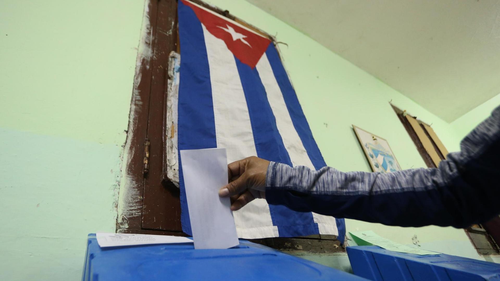 Das Bild zeigt nur den Arm der Frau, die den Stimmzettel gerade in den Schlitz einer blauen Urne steckt. An der lindgrünen Wand hängt eine kubanische Flagge. Das Bild zeigt nur den Arm der Frau, die den Stimmzettel gerade in den Schlitz einer blauen Urne steckt. An der lindgrünen Wand hängt eine kubanische Flagge.