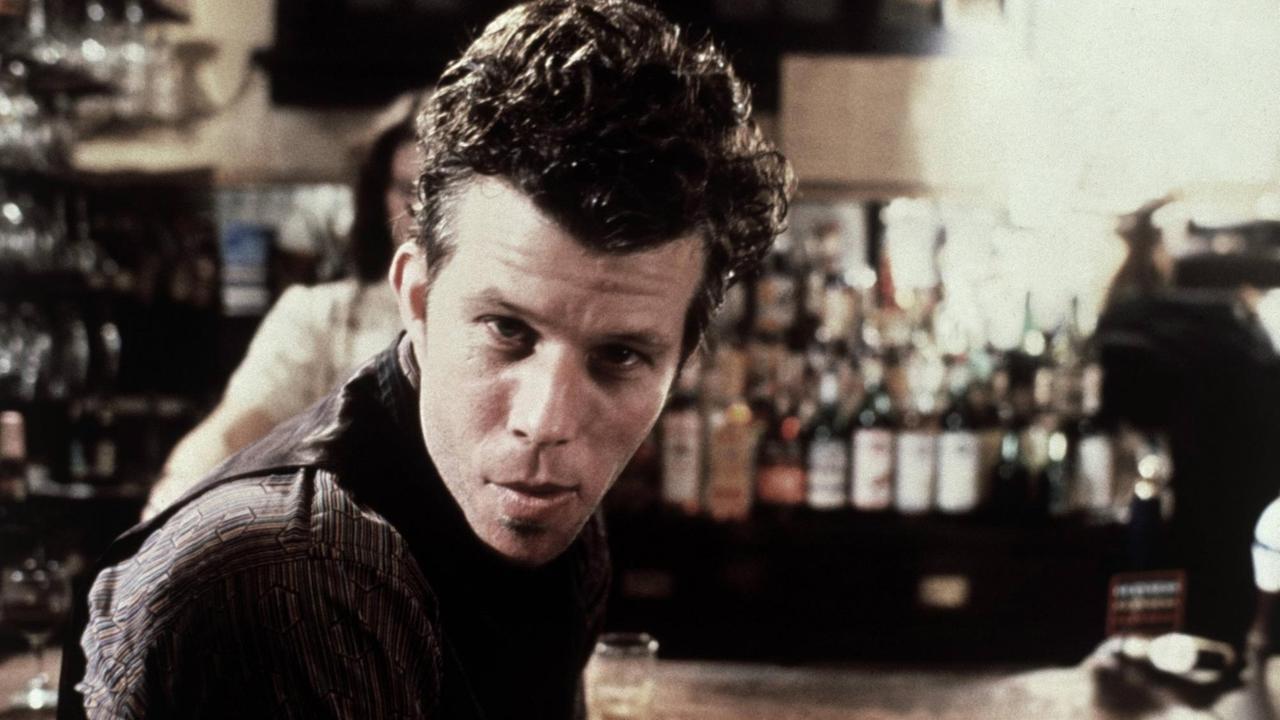 Tom Waits sitzt auf einem Hocker vor einer Bar und schaut direkt in die Kamera. Die Szene stammt aus dem Film "Wolfen" von 1981.