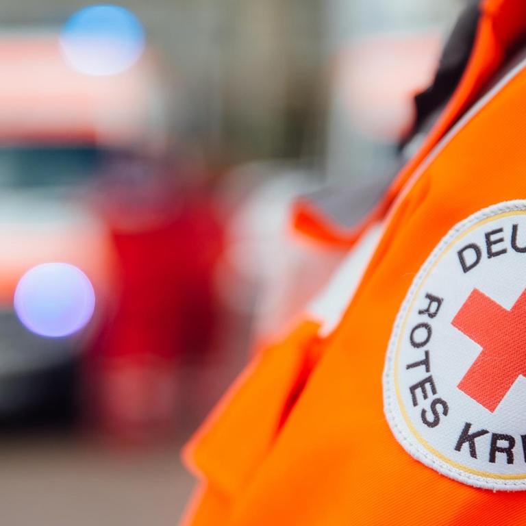 Ein Arm mit einem Aufnäher mit der Aufschrift "Deutsches Rotes Kreuz" ist vor einem Rettungsfahrzeug zu sehen.